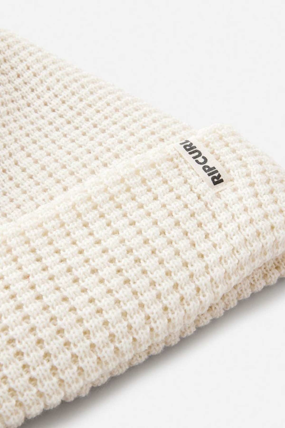 Close-up of the Rip Curl Anoeta Reg Pom Pom Beanie in cream, featuring a waffle-knit design, folded edge, small "RIP CURL" tag, and made from recycled polyester. Shown against a plain light grey background.