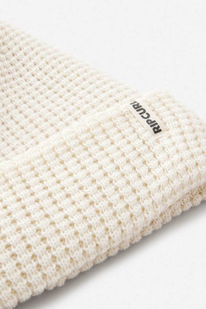 Close-up of the Rip Curl Anoeta Reg Pom Pom Beanie in cream, featuring a waffle-knit design, folded edge, small "RIP CURL" tag, and made from recycled polyester. Shown against a plain light grey background.