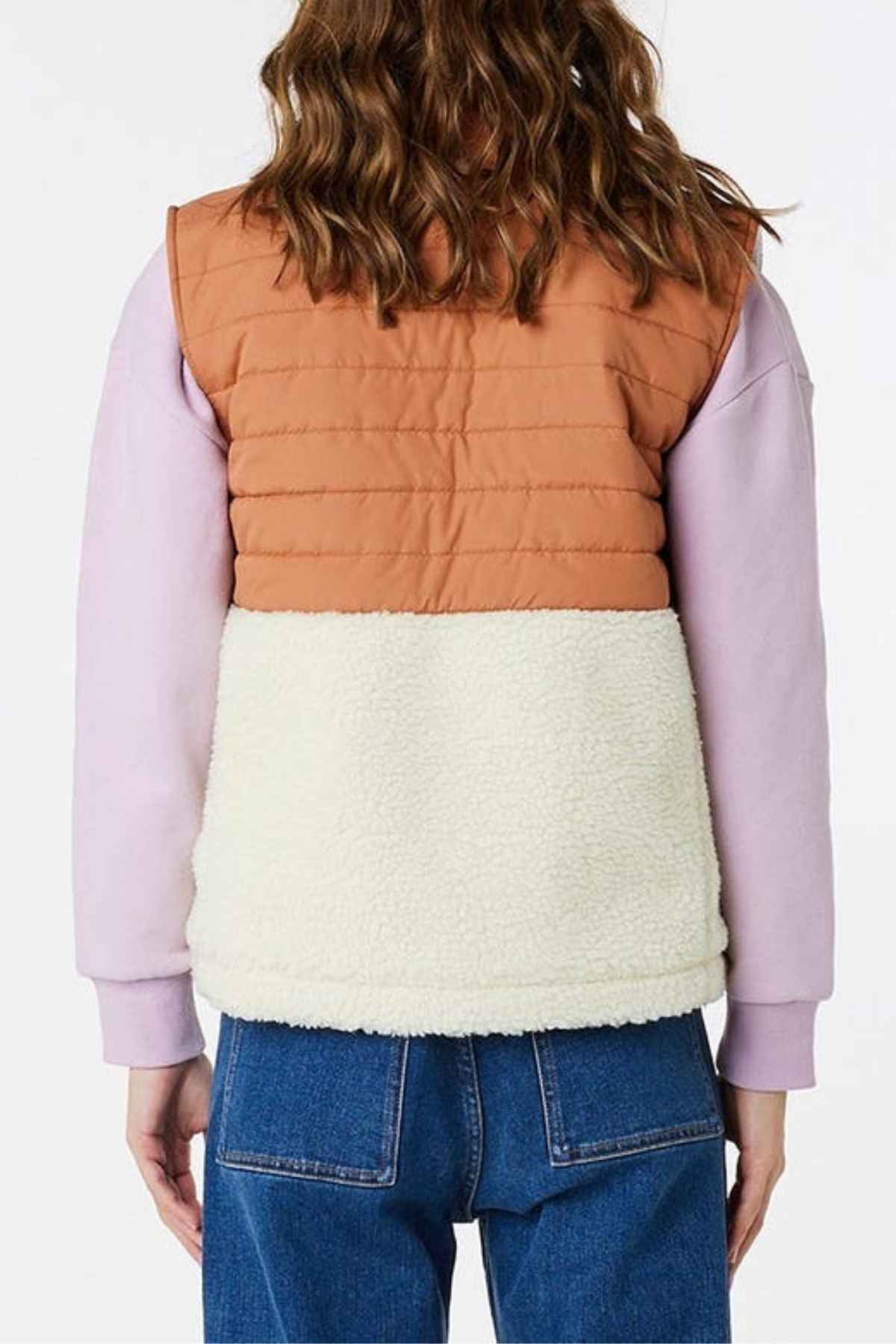A person with wavy brown hair wears the Rip Curl Anti-Series Anoeta Fleece Vest, which features a light brown quilted top and shower-resistant white fleece bottom. The sleeves are pastel pink, and they are in blue jeans against a plain white background.