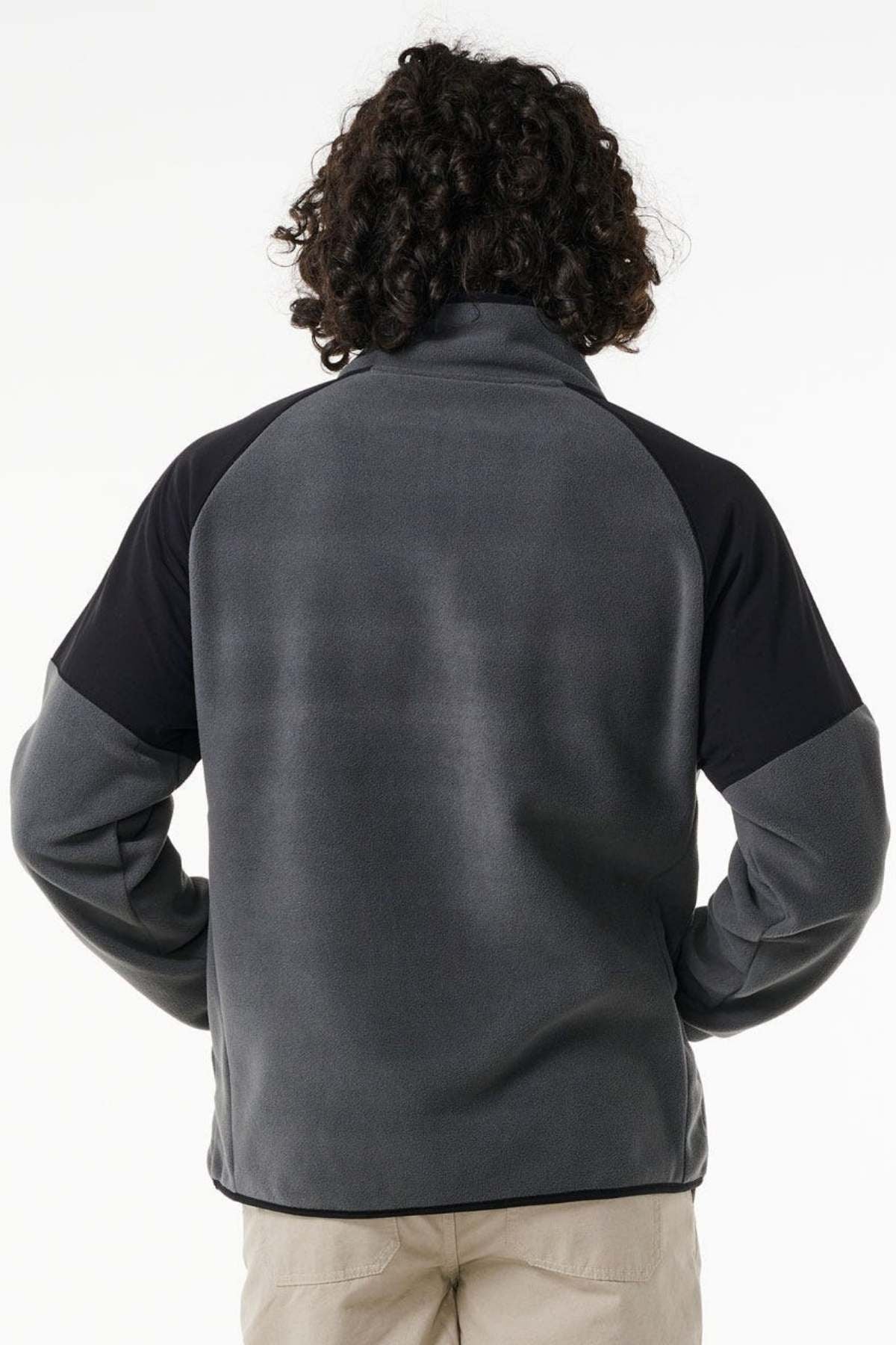 A person with curly hair is shown from the back wearing a dark gray and black Rip Curl Anti Series Archive Zip Crew and light-colored pants, standing against a plain white background.