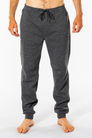 Barefoot against a white background, a person wears dark gray Rip Curl Departed Anti Series Trackpants crafted from bonded polar fleece. The relaxed fit features elastic cuffs and a drawstring. Their arms are slightly extended at their sides.