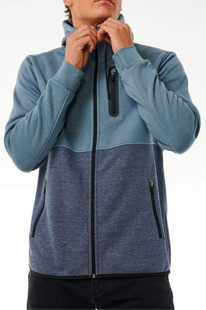 A person is seen adjusting the collar of a Rip Curl Anti Series Departed Zip Thru, a durable, water-resistant two-toned blue zip-up hoodie with three zippered pockets. The individual's face is hidden against a plain white background.