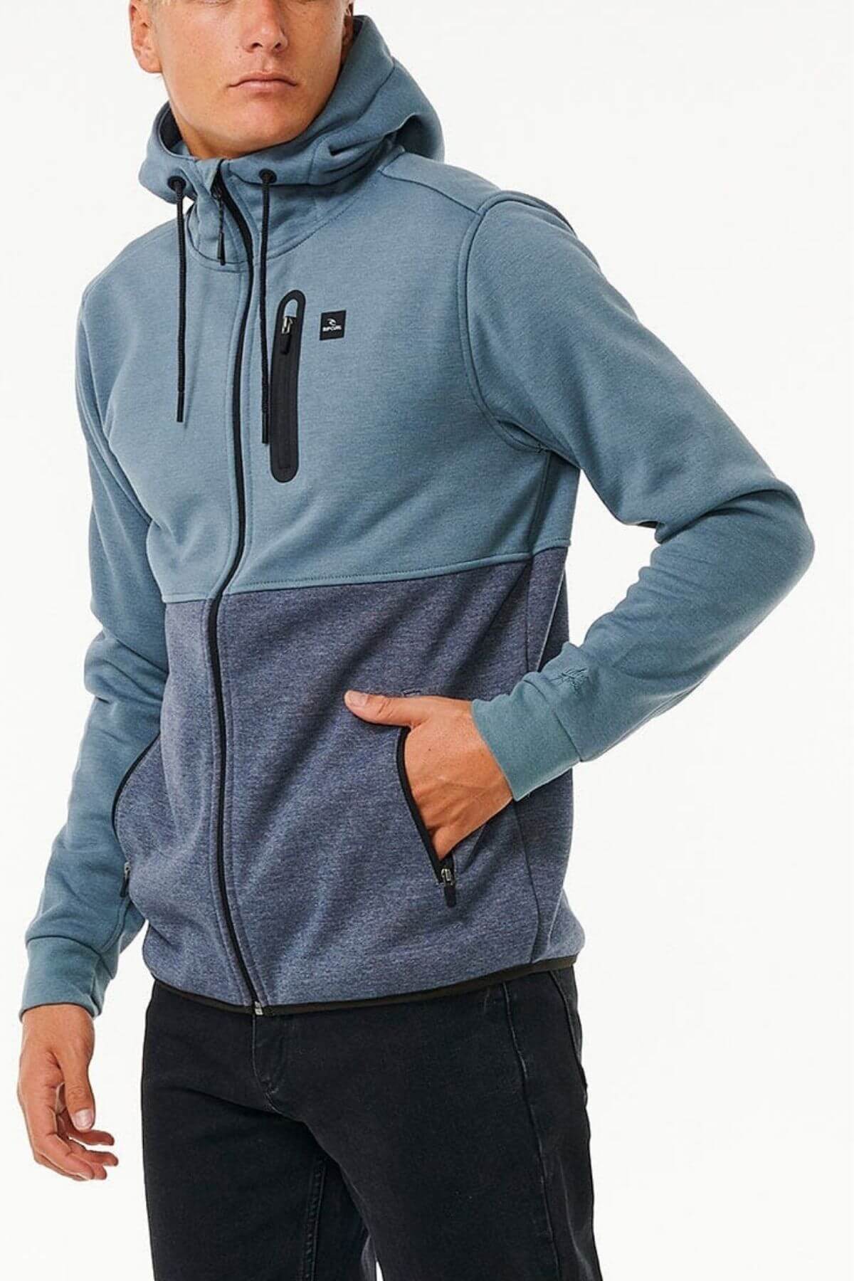 Wearing the Rip Curl Anti Series Departed Zip Thru, a person stands against a plain background. The two-toned blue hoodie with black zipper and chest pocket is crafted from durable water-resistant fabric for winter, featuring a drawstring hood and side pockets, paired perfectly with black pants.