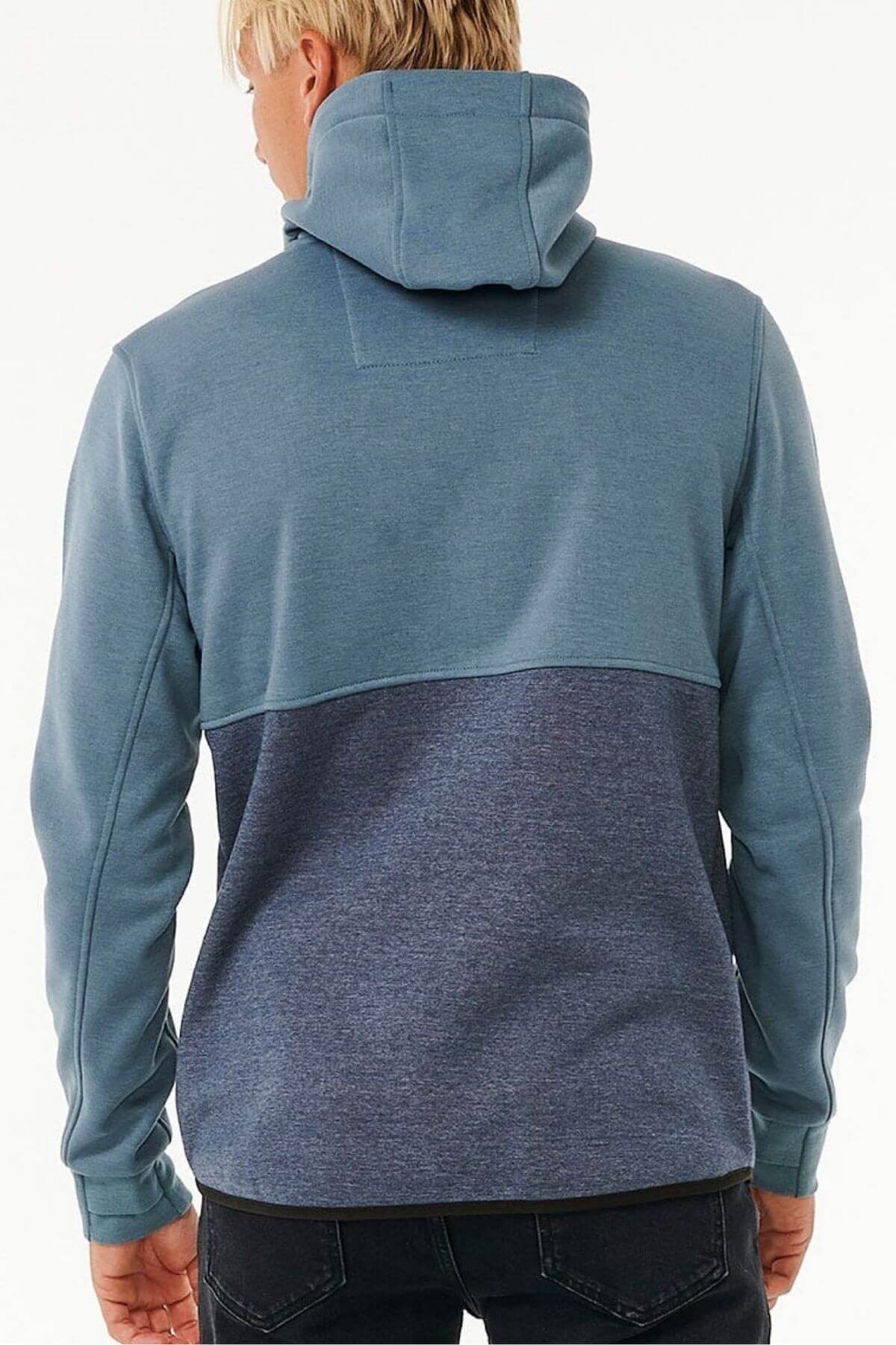 A person with blond hair, hood up, faces away wearing the Rip Curl Anti Series Departed Zip Thru. This two-toned blue hoodie has a lighter upper and darker lower half, paired with black pants—ideal for durable water-resistant winter wear.