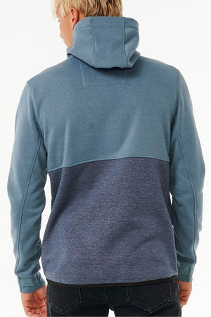 A person with blond hair, hood up, faces away wearing the Rip Curl Anti Series Departed Zip Thru. This two-toned blue hoodie has a lighter upper and darker lower half, paired with black pants—ideal for durable water-resistant winter wear.