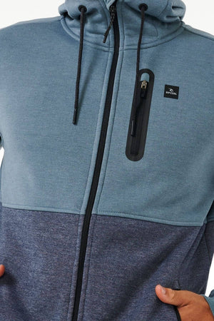 A close-up shows a person in a Rip Curl Anti Series Departed Zip Thru hoodie, two-toned blue with a small chest logo. This water-resistant winter piece features black zippers and drawstrings, while the person tucks their hands warmly into the pockets.