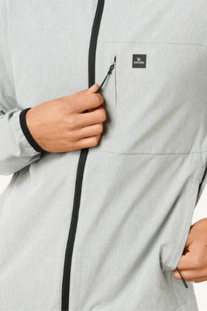 A person wearing the Rip Curl Anti Series Elite Jacket in light gray stretch polyester is unzipping the chest pocket with their right hand. The water-repellent jacket has black zipper accents and a small black logo on the chest.