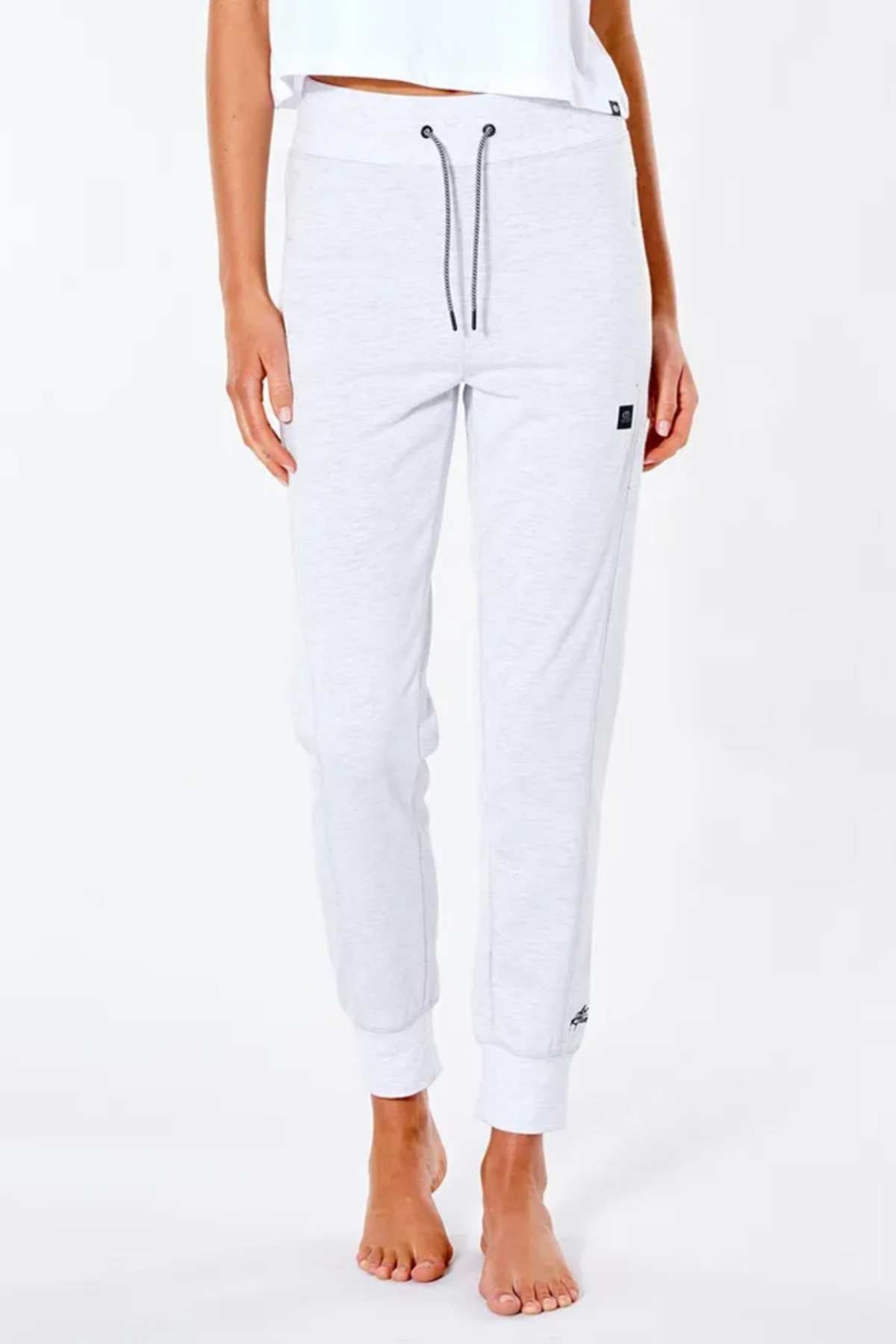 A person stands barefoot on a white background wearing Rip Curl Anti Series Flux II Trackpants in light gray. These water-repellent joggers have ribbed cuffs, a drawstring waist, and small black tags on the upper left thigh and lower right leg.