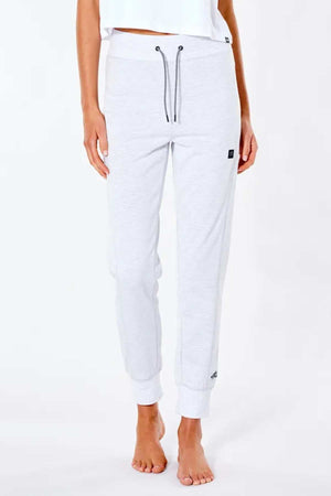 A person stands barefoot on a white background wearing Rip Curl Anti Series Flux II Trackpants in light gray. These water-repellent joggers have ribbed cuffs, a drawstring waist, and small black tags on the upper left thigh and lower right leg.