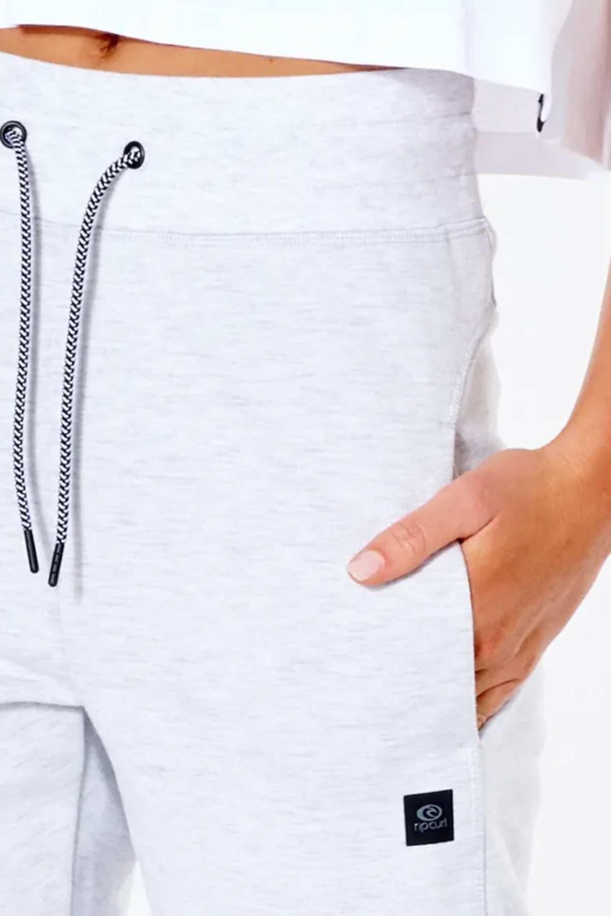 A person wears Rip Curl Anti Series Flux II Trackpant in light gray with a black drawstring, hand in right pocket. The trackpants have a small black logo tag on the side, and part of a white top is visible above the waist.
