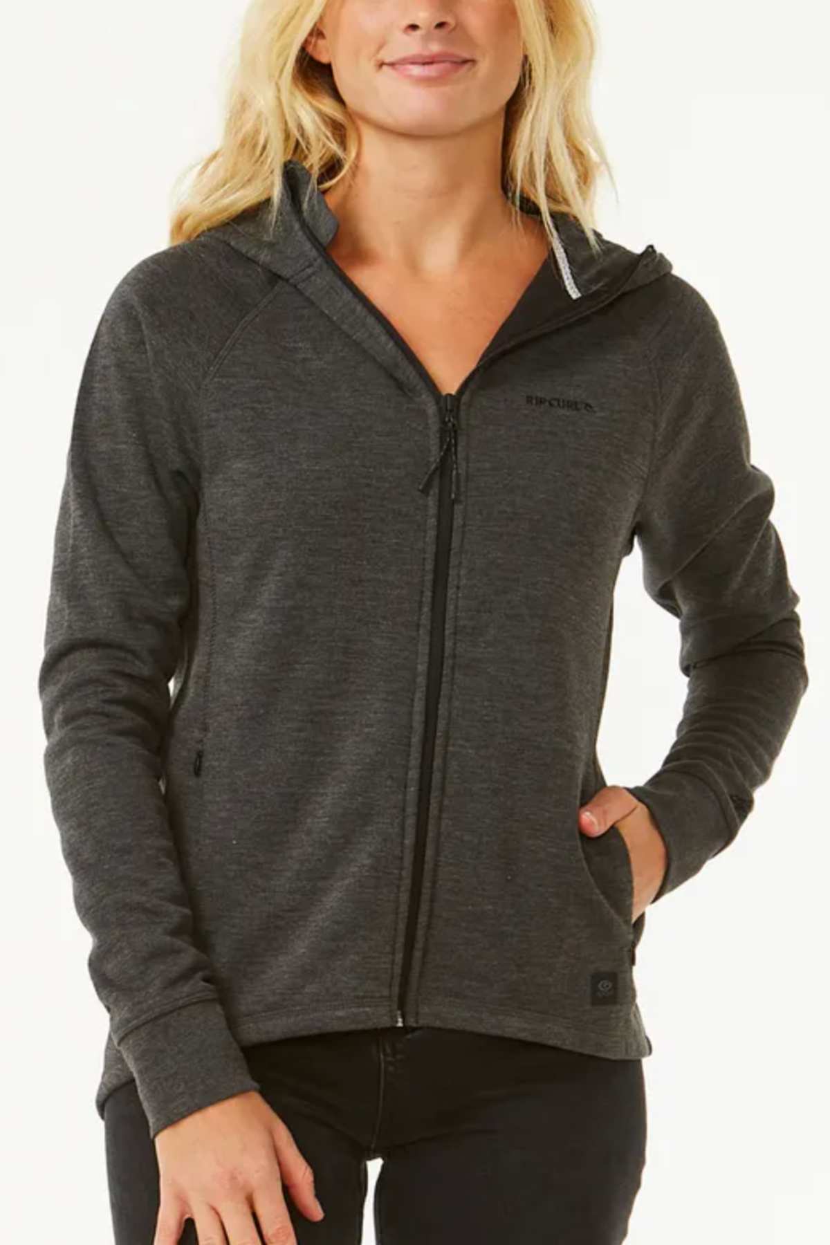 A person with long blonde hair wears a dark gray Rip Curl Anti Series Flux III Zip hooded jacket, featuring bonded polar fleece, long sleeves, and a subtle logo on the upper left side, with their left hand in the pocket.