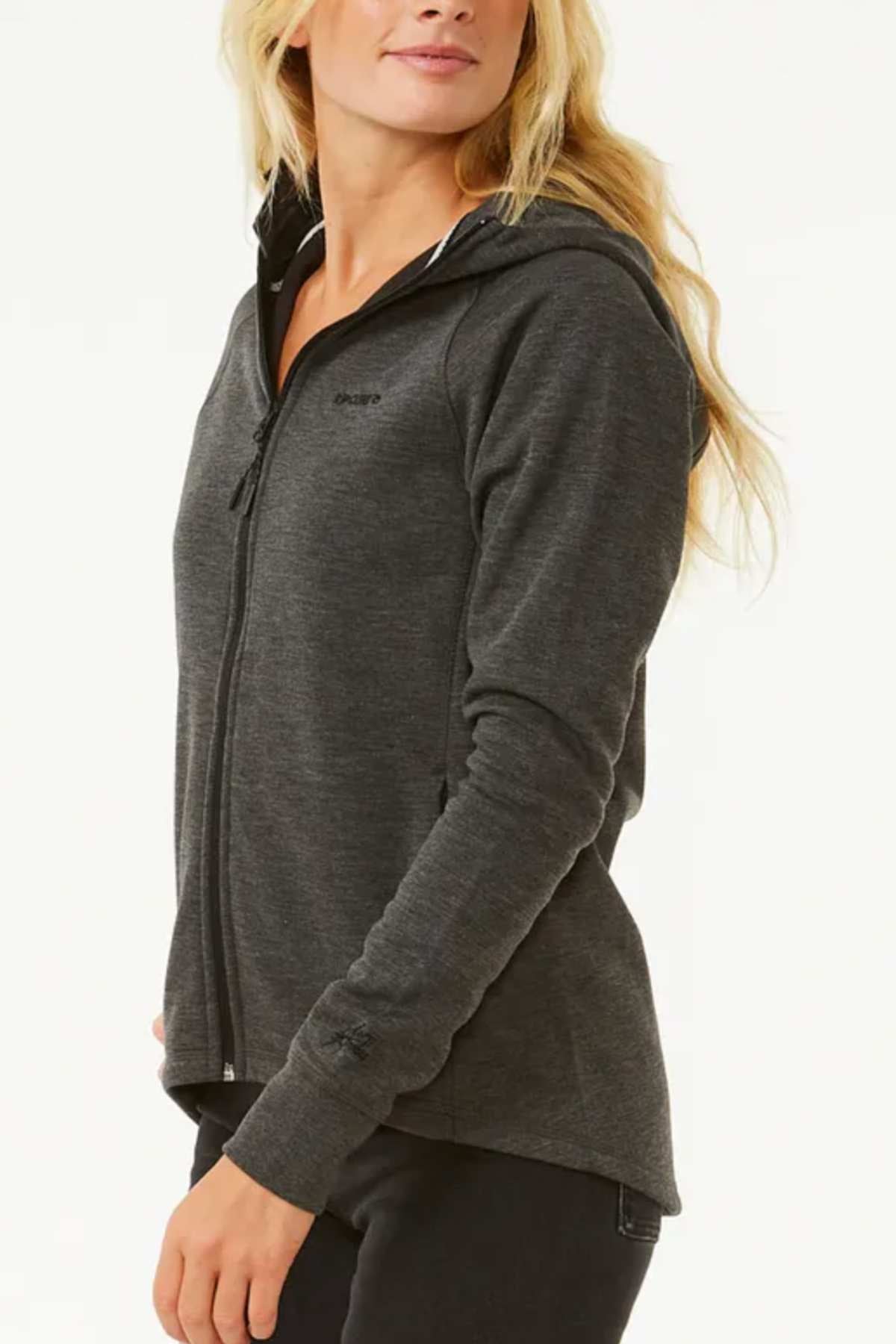 A person with long blonde hair in a dark gray Rip Curl Anti Series Flux III Zip, made from bonded polar fleece with DWR water repellent finish, stands against a light background, turning slightly to the side to display its fit.