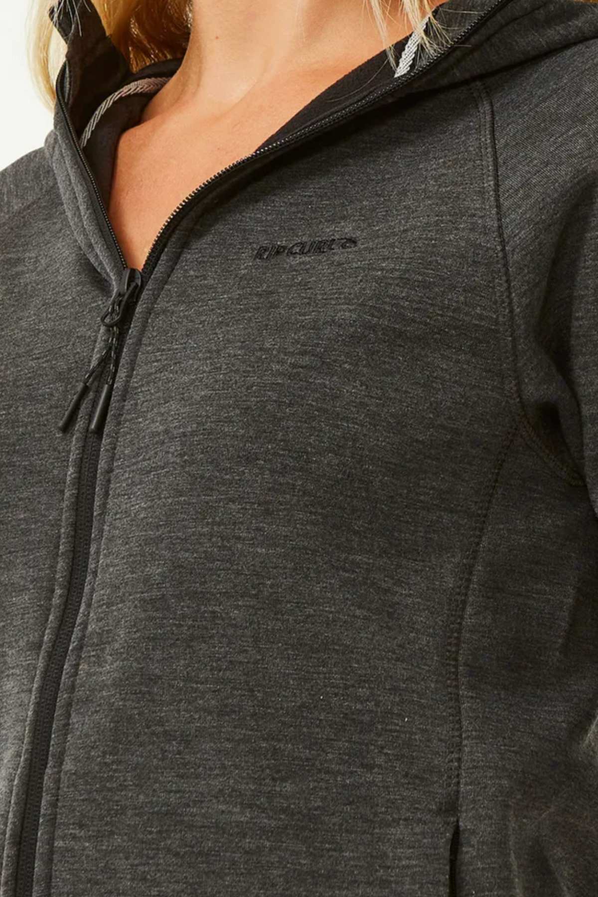 Someone is wearing a Rip Curl Anti Series Flux III Zip, a dark gray zip-up hoodie with bonded polar fleece lining, a subtle logo on the left chest, and DWR water repellent treatment highlighting the hood and zipper details.