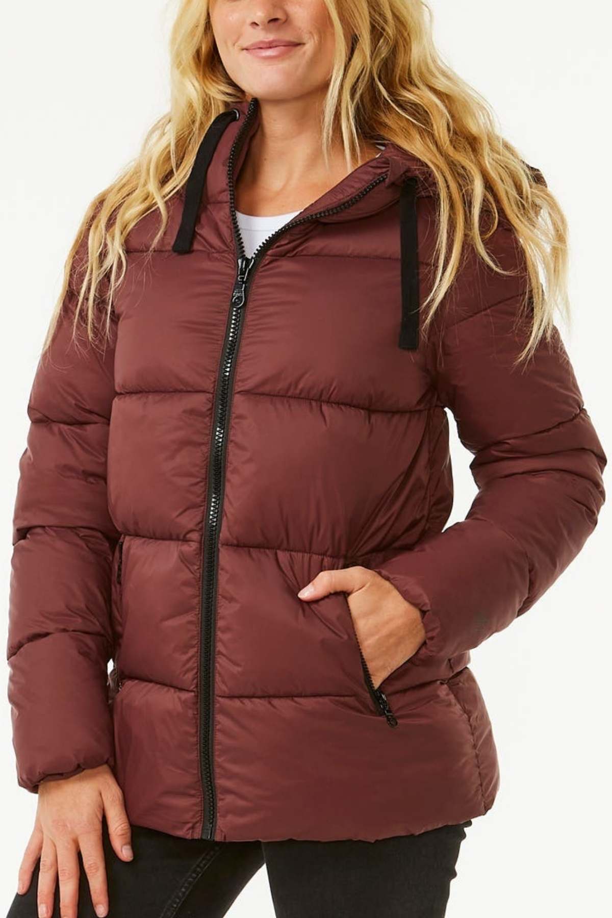 A woman stands against a plain white background, wearing a maroon Rip Curl Anti Series Insulated Jacket. The quilted design of the jacket complements her long, wavy blonde hair. She has one hand in her pocket and the jacket's front zipper and hood are clearly visible, adding to its stylish functionality. The jacket also features DWR water repellent treatment for added protection.