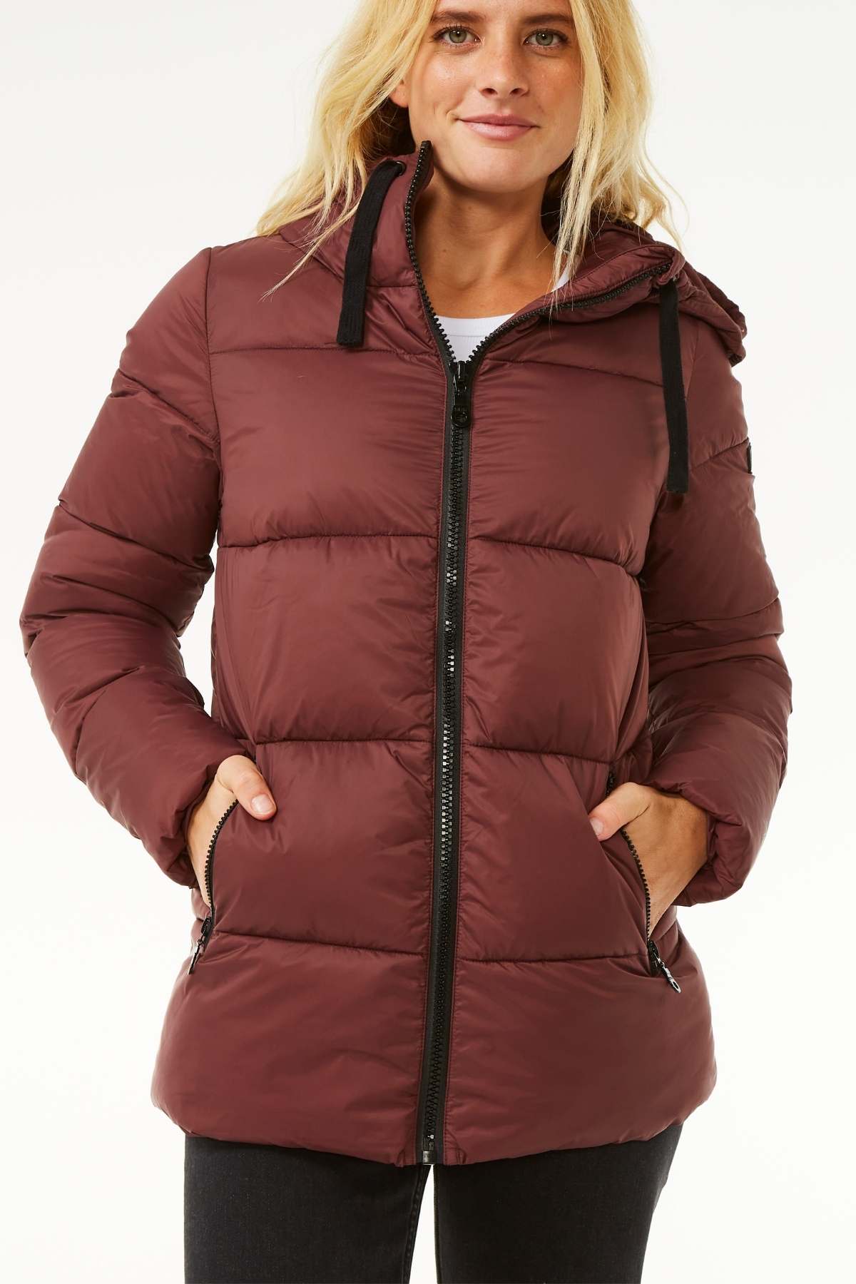 A person with long blonde hair is wearing a maroon Rip Curl Anti Series Insulated Jacket, which showcases a quilted design, hood, and black zipper. With their hands tucked into the jacket's pockets, they are standing against a plain white background.