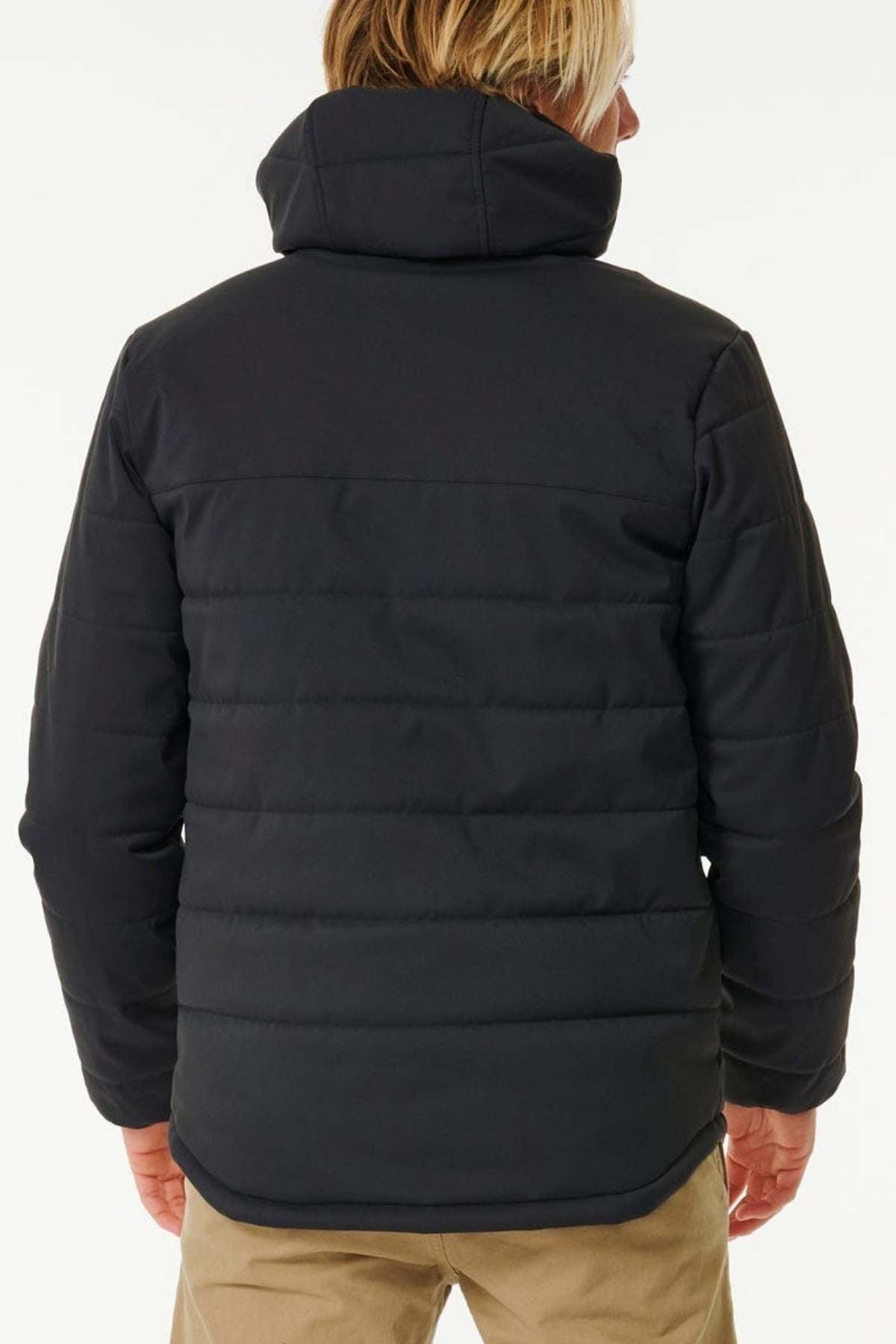 An outdoor enthusiast with short, light-colored hair is seen from behind, wearing a Rip Curl Anti Series Ridge Jacket paired with light-colored pants. The dark puffer jacket, known for its DWR water repellent qualities, stands out against the plain white background.