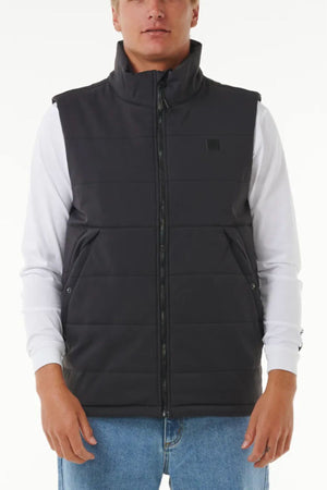 A man in a Rip Curl Anti Series Ridge Vest, featuring quilted padded lining, worn over a white long-sleeve shirt and blue jeans, stands against a plain white background.