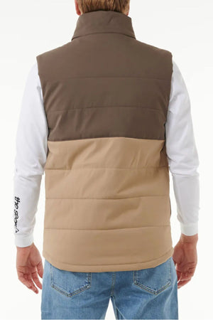 A person is shown from the back against a plain background wearing a brown and beige Rip Curl Anti Series Ridge Vest with quilted padded lining over a white long-sleeve shirt with writing on the sleeve and blue jeans.