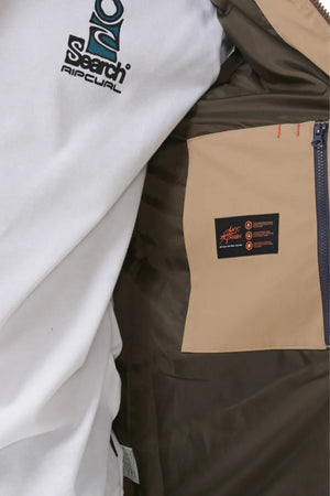 Close-up of someone wearing the Rip Curl Anti Series Ridge Vest, featuring a white shirt, brown jacket with quilted lining, beige pocket, blue zipper, and black "Heat Seeker" label—highlighting its premium comfort.