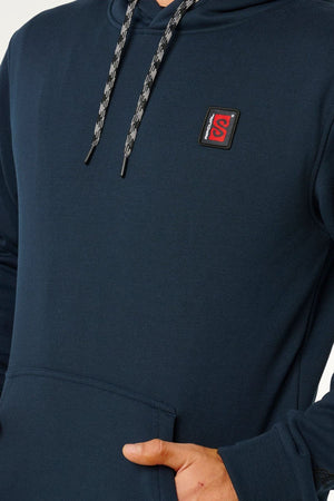 A person wearing a dark blue Rip Curl Anti Series Search Base Hood with a small red and black square logo on the chest and patterned drawstrings, shown from shoulders to hips, one hand partially in the front pocket.