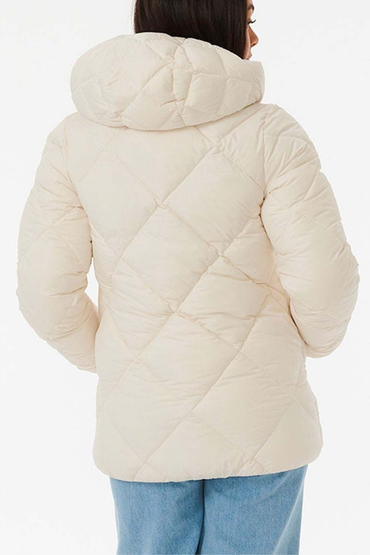 A person in a Rip Curl Anti Series Shorebreak Jacket, a cream-colored, water-repellent quilted puffer with a hood, stands with their back to the camera. Paired with blue jeans on a white background, it blends style and functionality for light shower resistance.