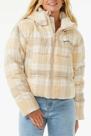 A person smiles against a plain white background wearing the Rip Curl Anti Series Surf Check Puffer—a beige and white plaid, shower-resistant jacket with a zipper, high collar, and "RIP CURL" on the chest—paired with light blue jeans.