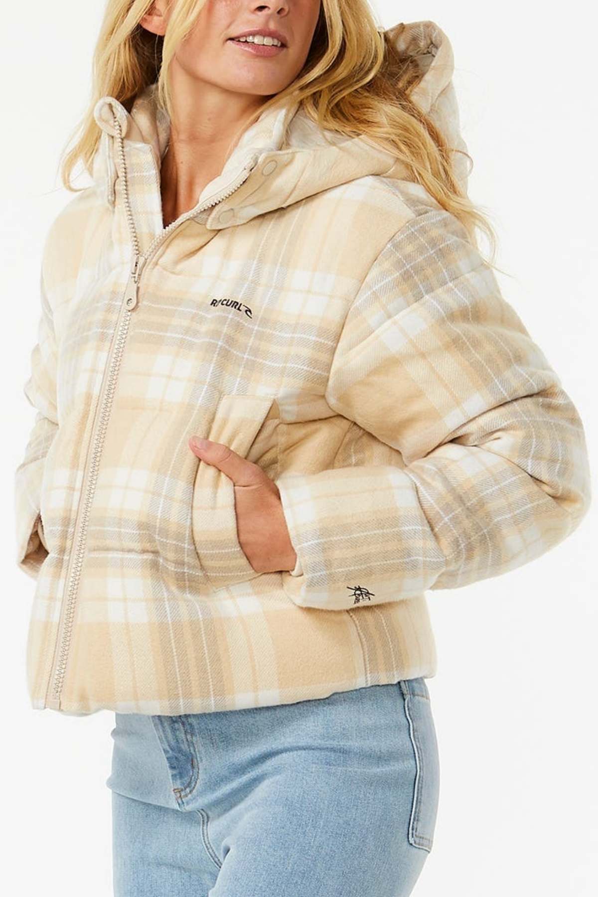 A woman wears a Rip Curl Anti Series Surf Check Puffer with shower resistance and light blue jeans, standing against a white background with her hands in the jacket pockets.