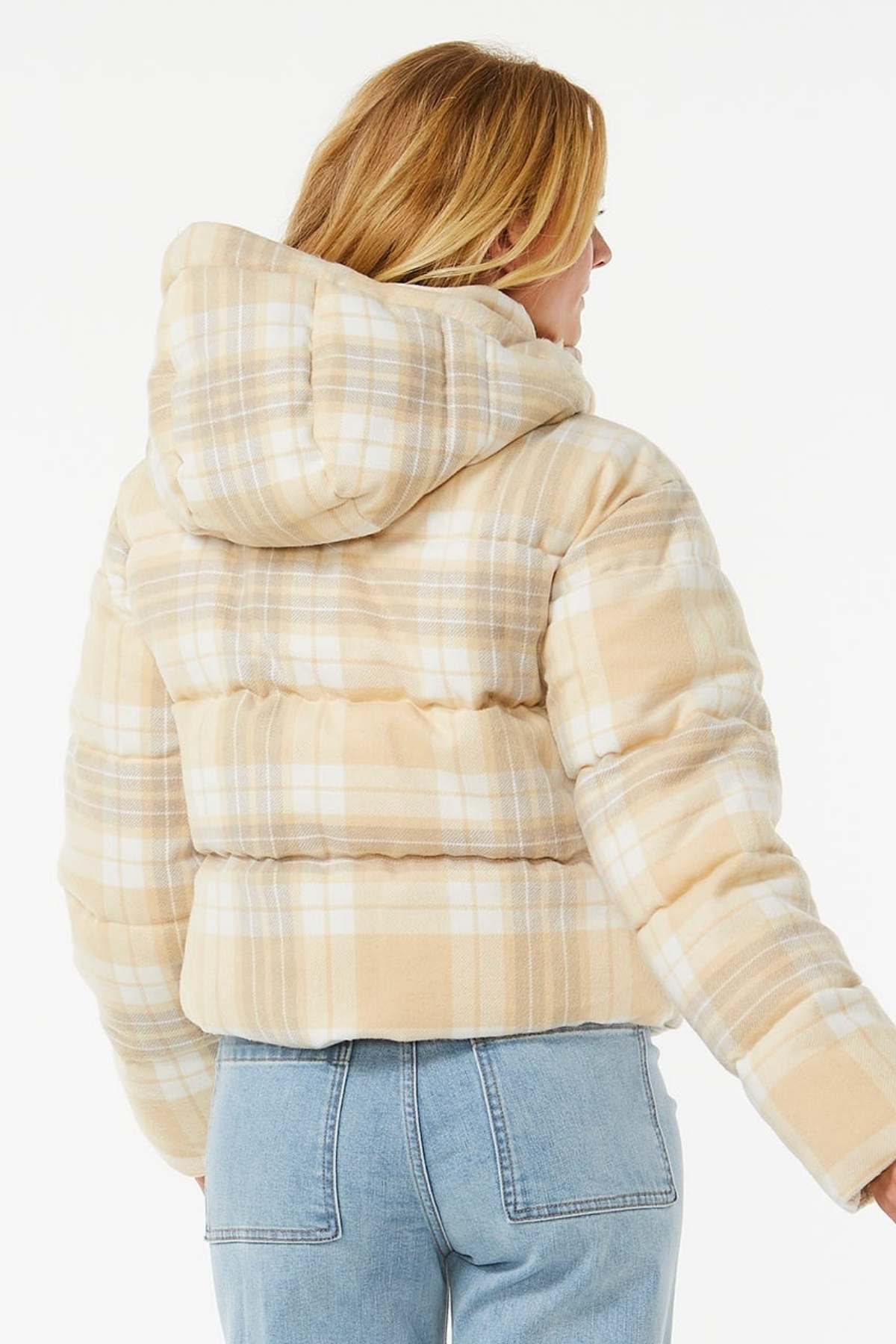 A person with blonde hair, seen from the back, wears a Rip Curl Anti Series Surf Check Puffer with a hood and shower resistance, paired with light blue jeans against a plain white background.