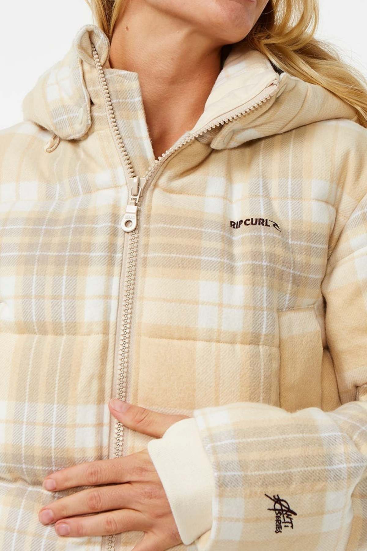 A person wears the Rip Curl Anti Series Surf Check Puffer, a beige and white plaid jacket with a hood, visible zippers, light blue accents, “RIP CURL” chest embroidery, and shower-resistant fabric. Their hand rests on the front.