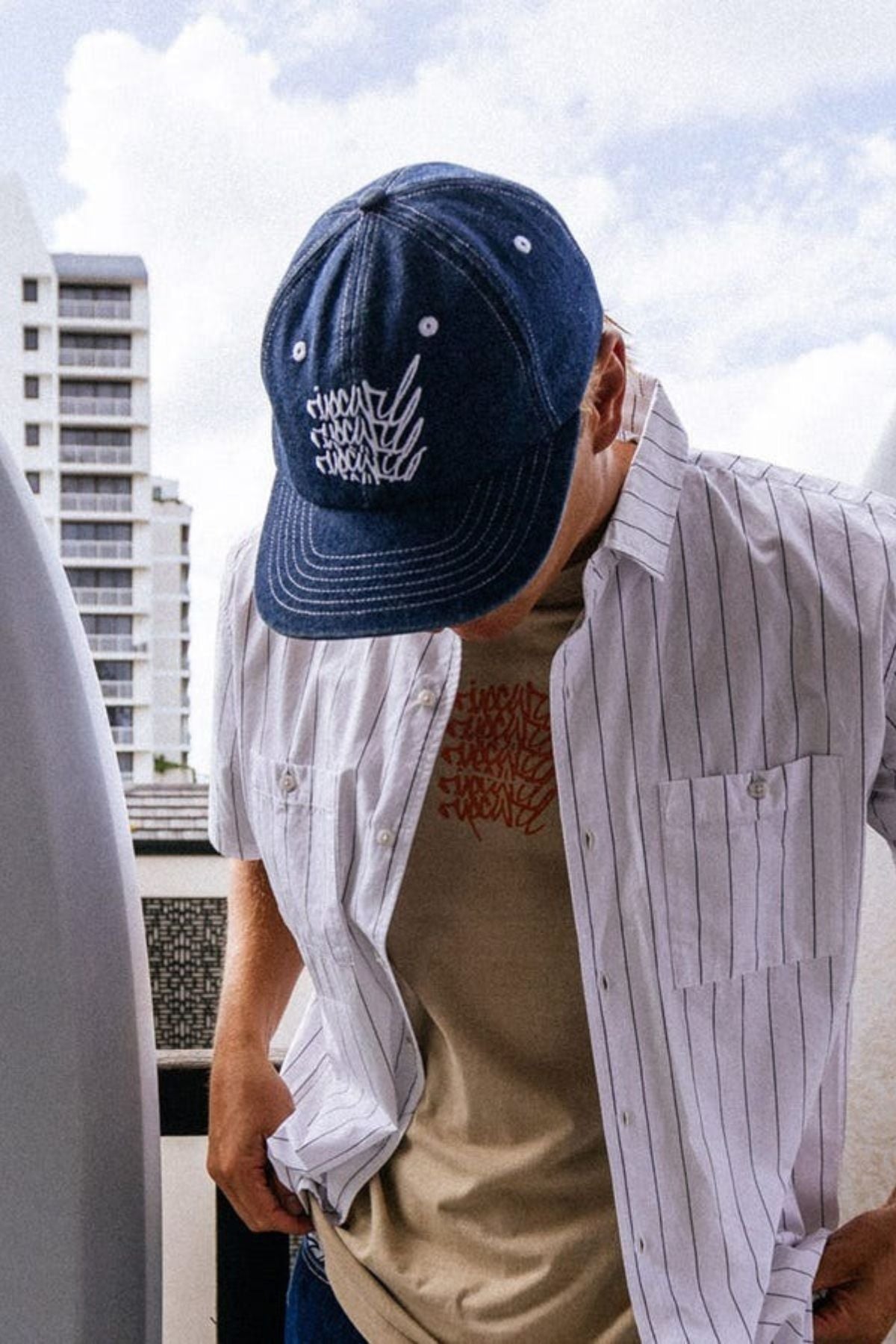 Wearing the Rip Curl Archive Denim SB Cap, a tan graphic tee, and a white pinstriped shirt, a person stands outdoors with their head down while modern white buildings appear in the background.
