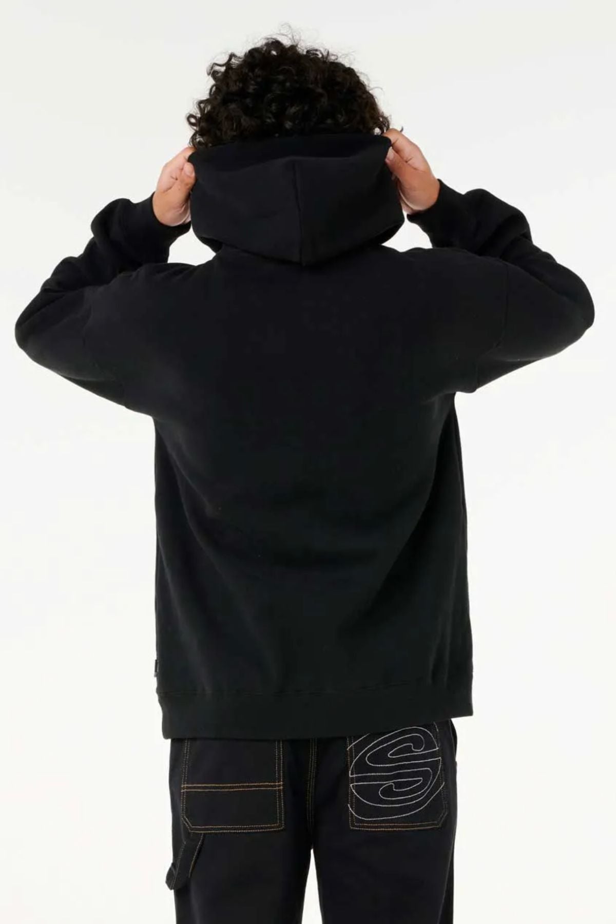 A person with curly hair, seen from behind, wears a loose-fitting Rip Curl Archive Hood and dark jeans with a large "S" stitched on the back pocket, holding the hood with both hands.