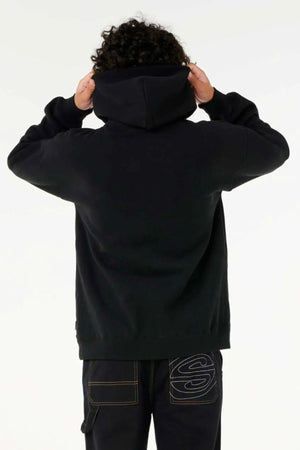 A person with curly hair, seen from behind, wears a loose-fitting Rip Curl Archive Hood and dark jeans with a large "S" stitched on the back pocket, holding the hood with both hands.