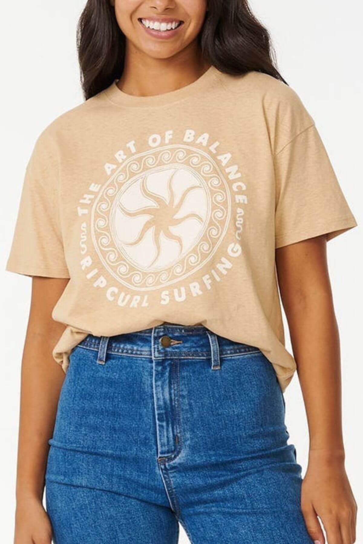 A person smiles in a Rip Curl Balance Relaxed Tee, showcasing "THE ART OF BALANCE" and "RIP CURL SURFING" around a wave and sun design on the front. This True Hemp beige tee pairs effortlessly with blue jeans for a stylish look.