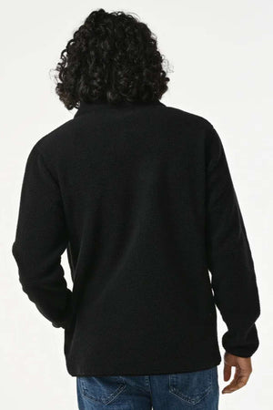 A person with curly hair is seen from the back, wearing a black Rip Curl Bells Polar Fleece jacket and blue jeans against a plain background.