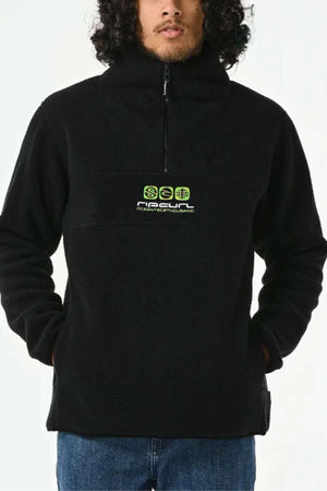 Someone wearing the Rip Curl Bells Polar Fleece, a black 1/4 zip fleece jacket with neon green chest text made from cozy Sherpa materials. The jacket highlights their curly hair. They pair it with blue jeans, hands in pockets, set against a plain white background.