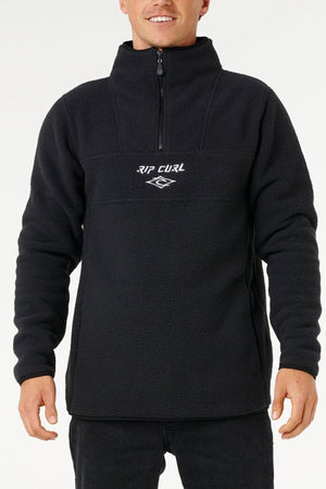 A person in a black Rip Curl Bells Polar Fleece with a front half-zip, zip pockets, and white "RIP CURL" chest embroidery stands against a plain white background.