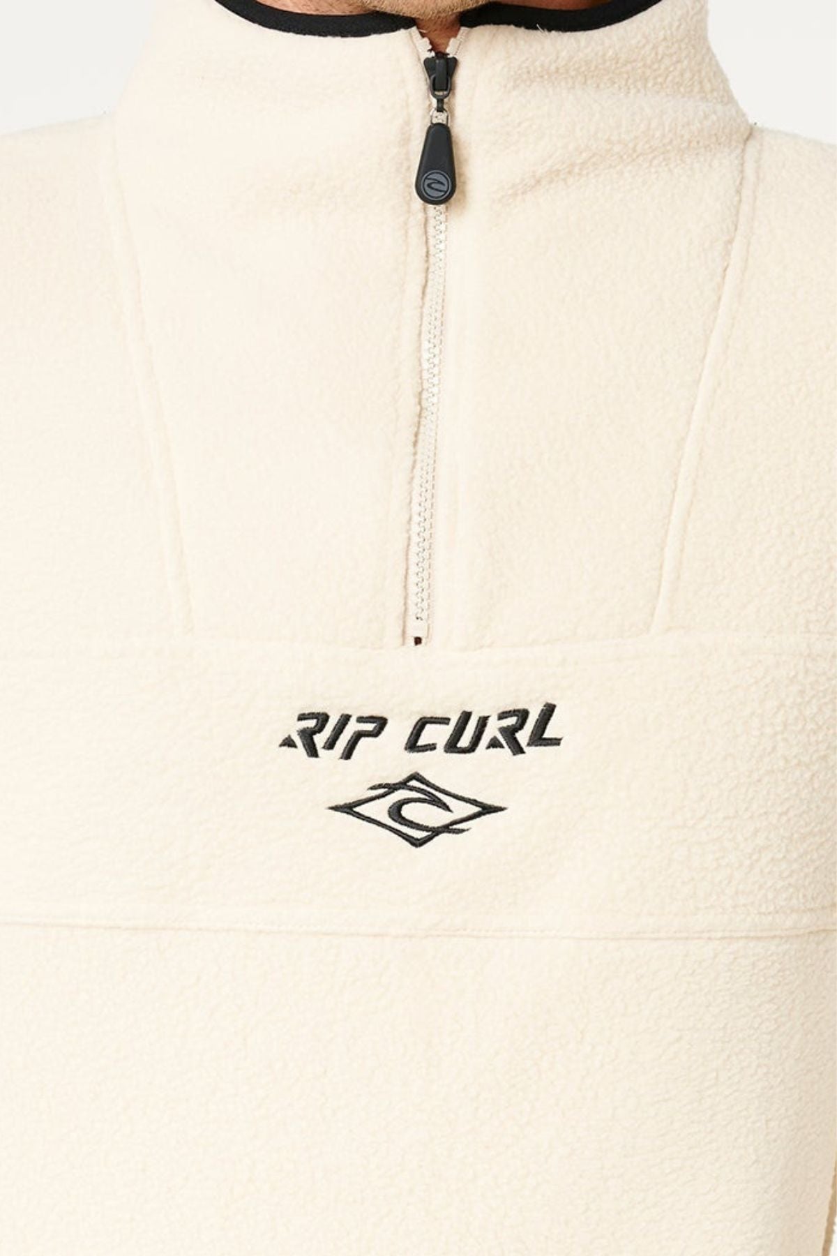 Close-up of a person wearing the cream Rip Curl Bells Polar Fleece pullover with a half-zip and embroidered "RIP CURL" logo. The iconic black text and wave symbol stand out on the soft polar fleece; lower part of face visible only.