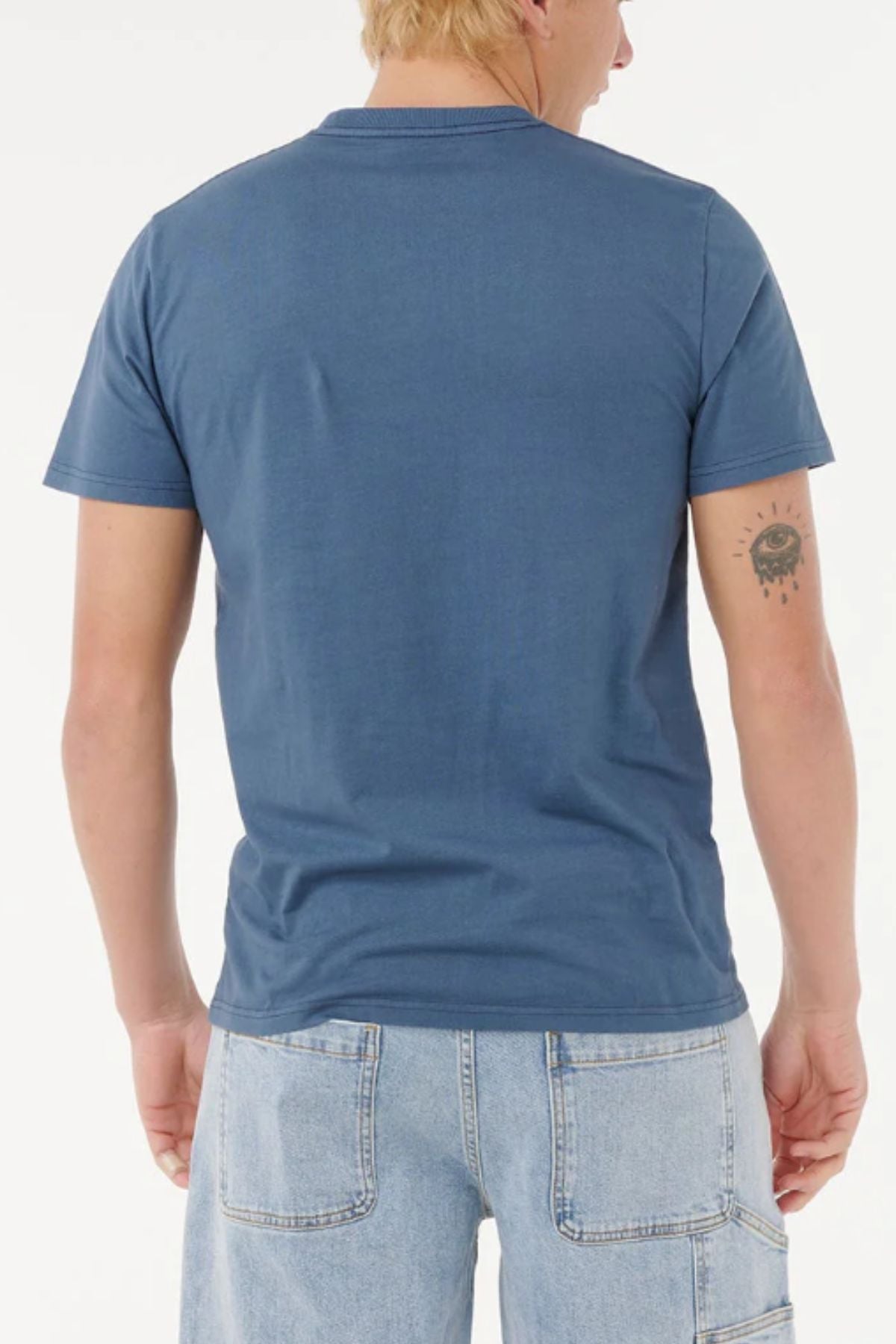 A person with short blond hair is seen from behind, wearing a blue Rip Curl Big Mumma Icon Tee and light blue denim shorts. A sun tattoo with a face is on the back of their left arm. The background is plain white.