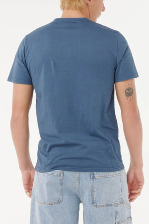A person with short blond hair is seen from behind, wearing a blue Rip Curl Big Mumma Icon Tee and light blue denim shorts. A sun tattoo with a face is on the back of their left arm. The background is plain white.