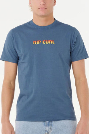 A person wears a blue Rip Curl Big Mumma Icon Tee with bold yellow and orange gradient lettering on the chest, paired with light blue jeans, against a plain white backdrop.