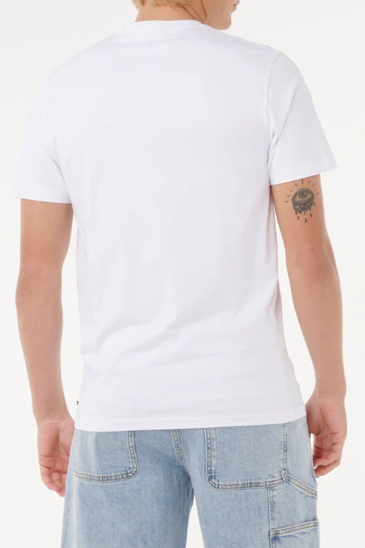 Person standing with their back to the camera, wearing a Rip Curl Big Mumma Icon Tee in plain white and light blue denim jeans. A tattoo of an eye with dripping details is visible on their upper right arm.