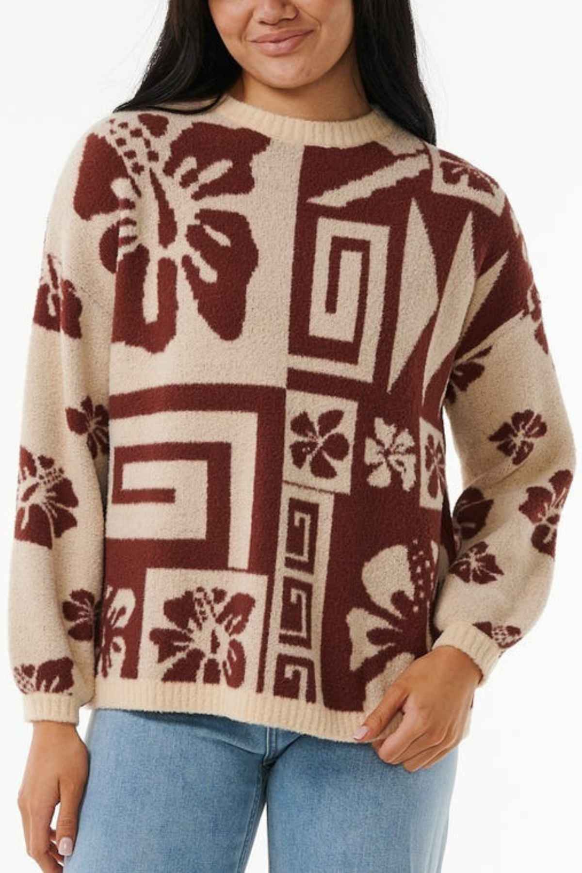 A person is wearing the Rip Curl Block Party Jacquard Crew, a beige jacquard knit sweater with an abstract red floral and geometric pattern. The sweater features a ribbed neckline, cuffs, and hem. They are also dressed in light blue jeans against a plain white background.