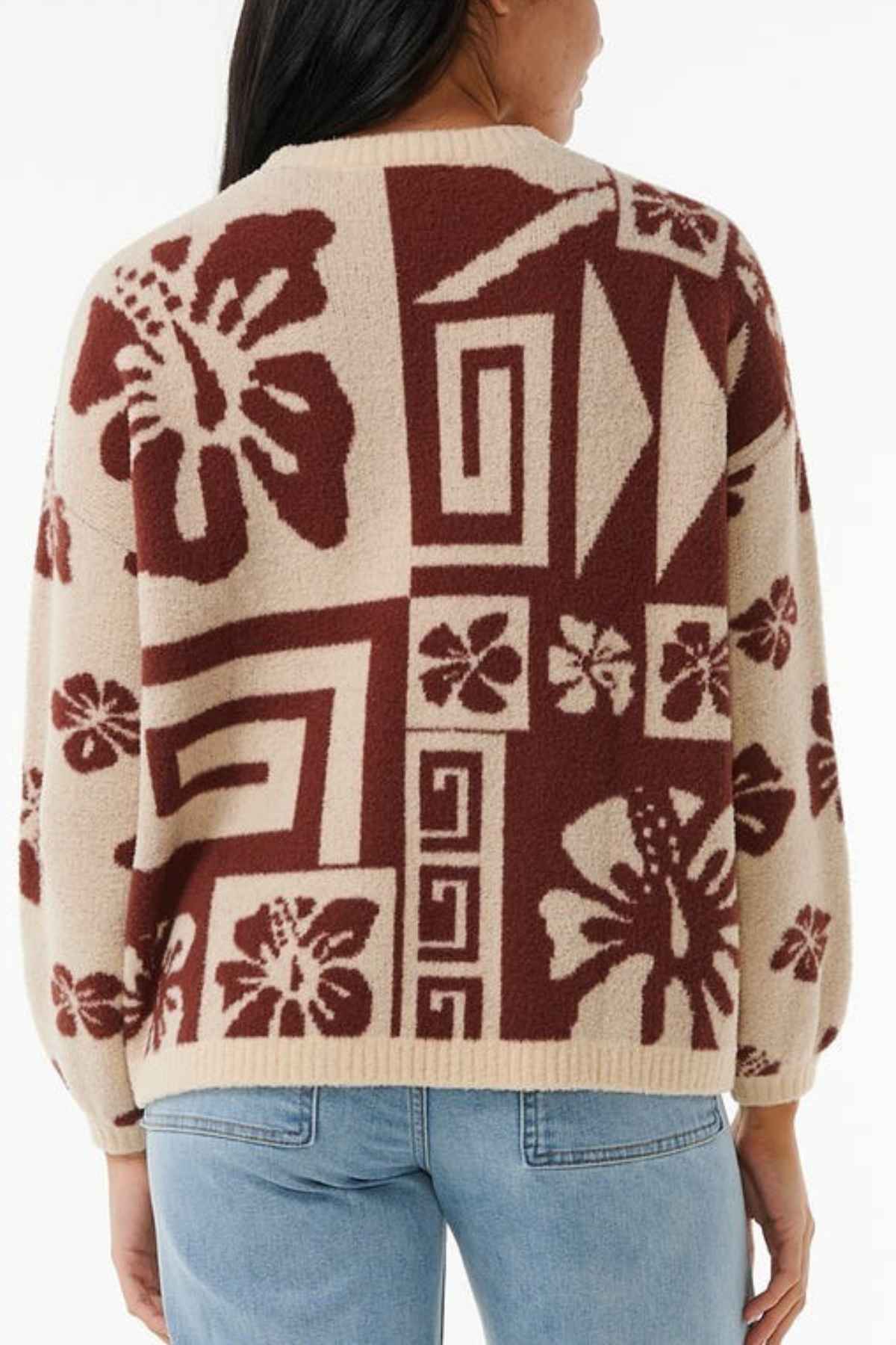 A person wearing the Rip Curl Block Party Jacquard Crew with a beige hue, featuring a striking brown geometric and floral pattern. The individual is turned away from the camera, highlighting the detailed jacquard knit of the sweater. Paired with this ensemble are light blue jeans.