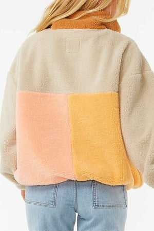 A person with long blonde hair wears a Rip Curl Block Party Polar Fleece in tan, peach, and yellow with light blue jeans, showing the back of the fleece and the upper part of the jeans.