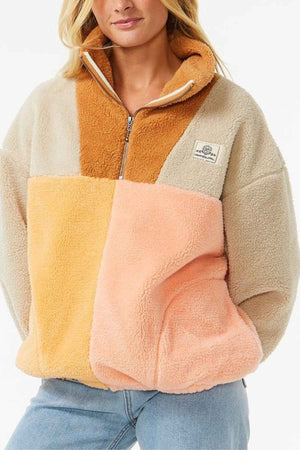 A woman wears the Rip Curl Block Party Polar Fleece in beige, tan, yellow, and peach with a half-zip collar and side pockets, styled with light blue jeans.