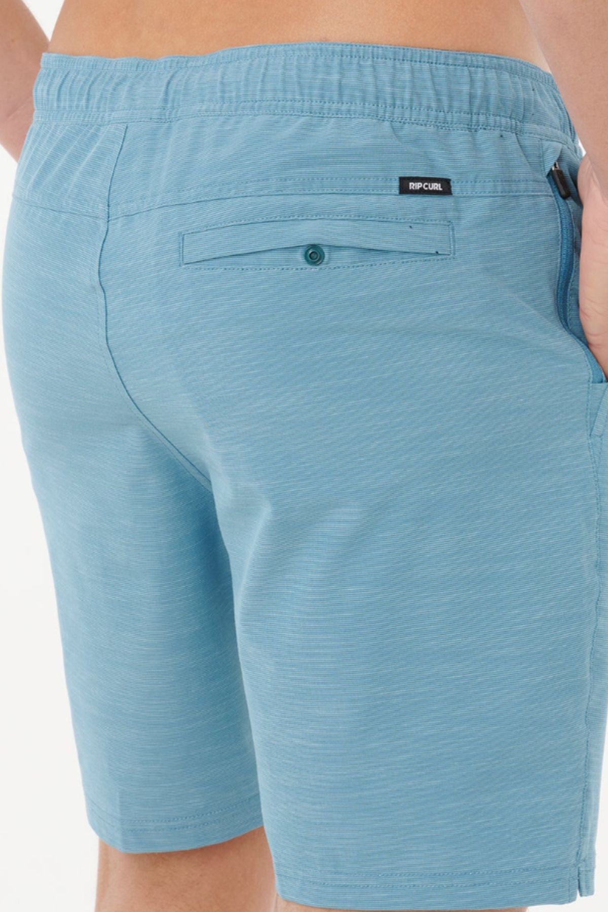 Close-up of a person wearing Rip Curl Boardwalk Jackson Volley shorts, highlighting the buttoned back pocket, small brand label near the waistband, and one hand partially tucked into a side pocket.