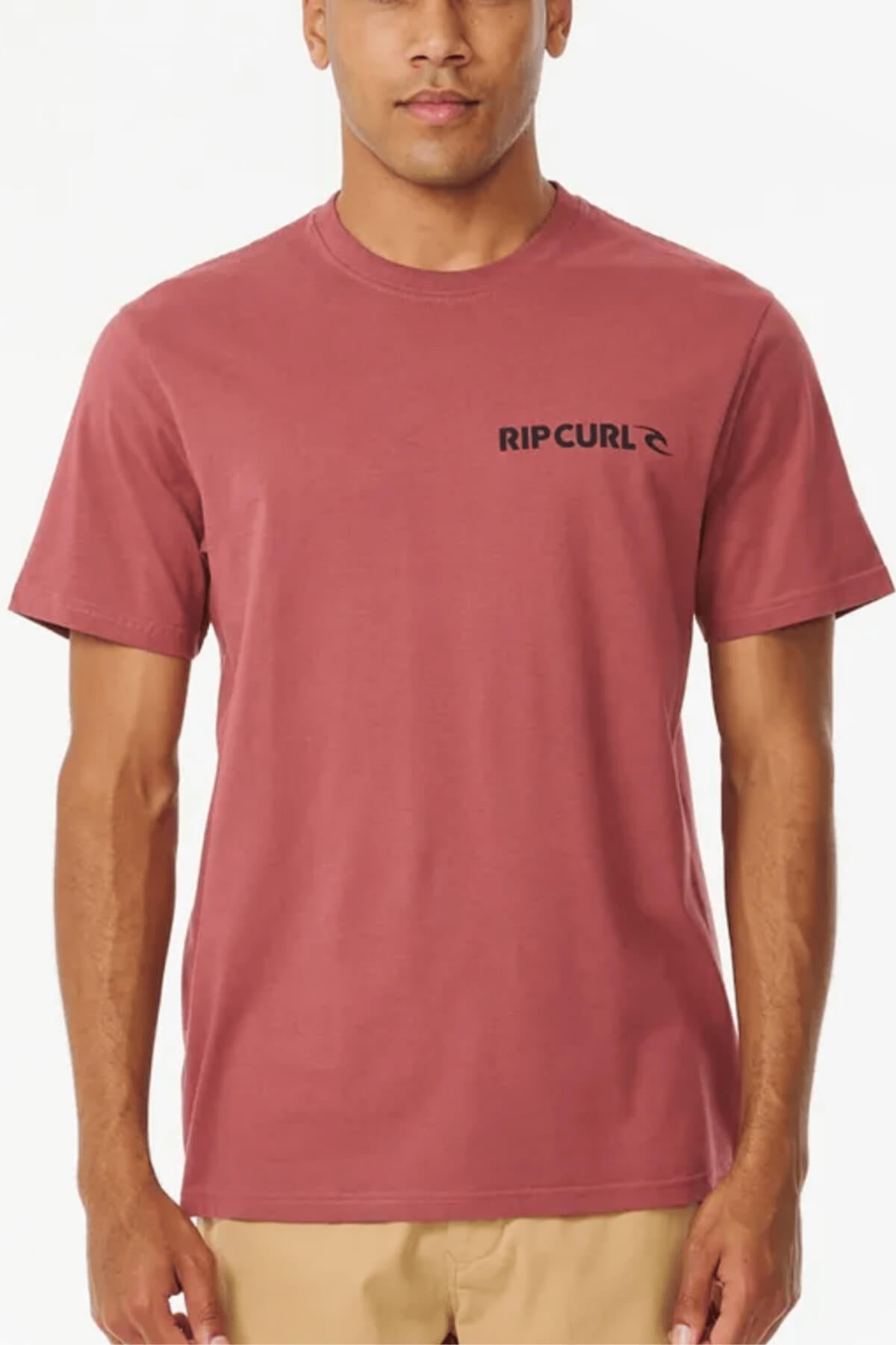A person is wearing a classic Rip Curl Brand Icon Tee in red-brown with beige pants. The 100% cotton t-shirt displays the black Rip Curl logo on the left chest. They stand against a plain white background.