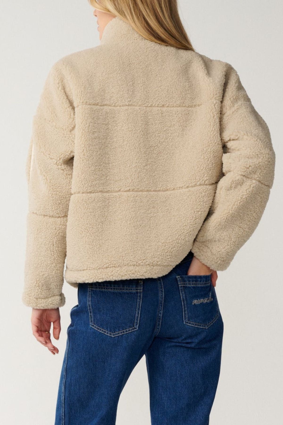 A person with long blonde hair is seen from the back against a plain light background, wearing blue jeans and a beige Rip Curl Cala Nights Sherpa Jacket featuring premium herringbone taping.