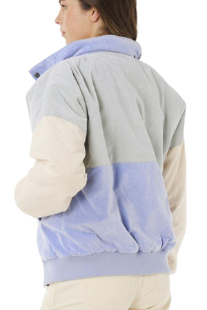 A person with shoulder-length brown hair is seen from the back wearing the Rip Curl Cala Vadella Jacket, a loose fit cotton corduroy jacket featuring retro color-blocking in pale blue, light gray, and beige with a ribbed hem.