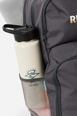 A light-colored water bottle with a black lid and "RUN" graphic sits in the mesh side pocket of a dark gray Rip Curl Chaser 33L Backpack, made from recycled polyester and compatible with 15" computers.